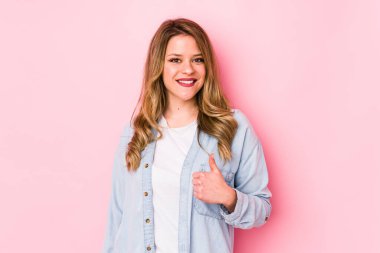 Pembe arka planda izole edilmiş genç beyaz kadın gülümsüyor ve başparmağını kaldırıyor.