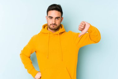 Mavi arka planda izole genç beyaz adam baş parmağını aşağı gösteriyor, hayal kırıklığı konsepti.