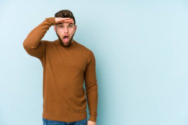 Kafkasyalı genç adam mavi arka planda izole edilmiş, uzaklara bakıyor ve elini alnına koyuyor..