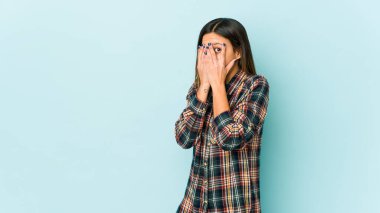 Mavi arka planda izole edilmiş genç bir kadın parmaklarının arasından korkmuş ve gergin bir şekilde göz kırpıyor..