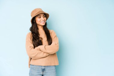 Mavi arka planda izole edilmiş bir şapka takan genç Hintli kadın kollarını kavuşturarak gülümsüyor..