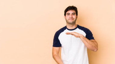 Bej arka planda izole edilmiş genç bir adam iki eliyle bir şey tutuyor, ürün sunumu.