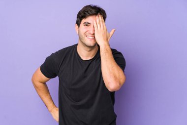 Mor arka planda izole edilmiş genç bir adam yüzünün yarısını avuç içiyle kaplayarak eğleniyor..