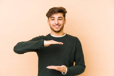 Bej arka planda izole edilmiş genç bir Arap adam iki eliyle bir şey tutuyor, ürün sunumu..