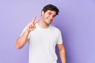 Mor arka planda izole edilmiş genç adam zafer işareti ve geniş bir gülümseme sergiliyor..