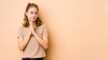 Beyaz tenli genç bir kadın bej arka planda izole edilmiş dua ediyor, bağlılık gösteriyor, dindar biri ilahi ilham arıyor..