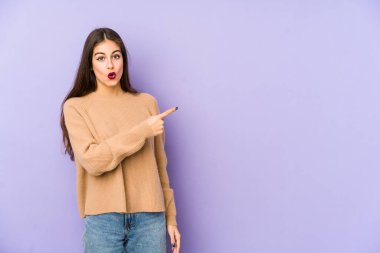 Mor arka planda izole edilmiş genç beyaz kadın gülümsüyor ve boş alanda bir şeyler gösteriyor..