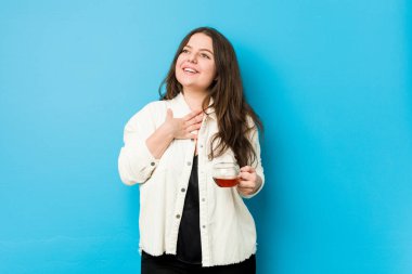 Genç kavisli kadın elinde çay fincanıyla kahkahalar atıyor elini göğsüne koyuyor..