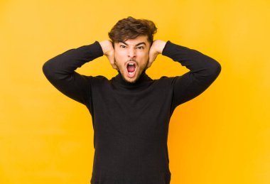 Sarı arka planda yalıtılmış genç bir Arap adam kulaklarını elleriyle kapatıyor ve çok fazla ses duymamaya çalışıyor..