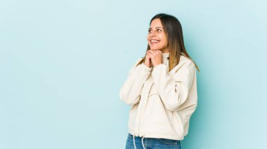 Mavi arka planda izole edilmiş genç bir kadın ellerini çenesinin altında tutuyor ve mutlu bir şekilde yan tarafa bakıyor..