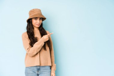 Mavi arka planda izole edilmiş bir şapka takan genç Hintli kadın gülümsüyor ve boş alanda bir şeyler gösteriyor..
