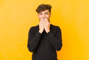 Sarı arka planda izole edilmiş genç bir Arap adam bir şeye gülüyor, elleriyle ağzını kapatıyor..