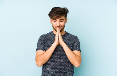 Mavi bir arka planda izole edilmiş genç bir Arap, el ele tutuşup, ağzının kenarında dua ediyor, kendinden emin..
