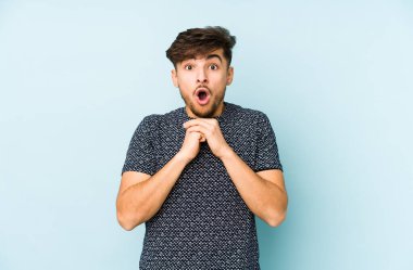 Genç Arap bir adam mavi bir arka planda izole edilmiş şans için dua ediyor, şaşırmış ve ön tarafa bakan ağzını açıyor..