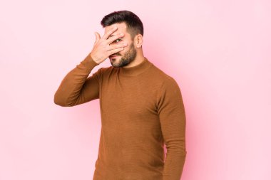 Pembe arka planda genç beyaz bir adam kameraya parmaklarıyla göz kırpıyor, yüzünü kapatmaktan utanıyor..