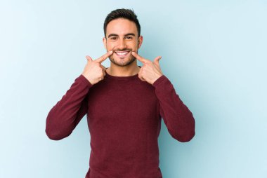 Mavi arka planda izole edilmiş genç beyaz adam gülümsüyor, parmaklarını ağıza doğrultuyor..