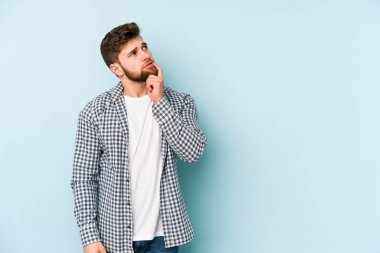 Mavi arka planda izole edilmiş genç beyaz adam şüpheli ve şüpheci bir ifadeyle yana bakıyor..