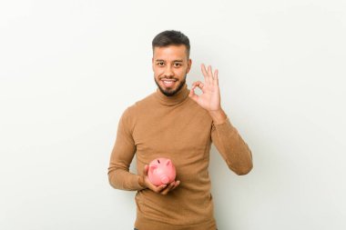 Genç Güney Asyalı bir adam elinde kumbarayla neşeli ve kendinden emin bir şekilde iyi niyet gösterisi yapıyor..