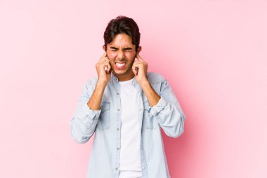 Pembe arka planda poz veren genç beyaz bir adam kulaklarını elleriyle kapatıyor..