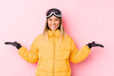 Pembe arka planda kayak kıyafeti giyen beyaz bir kadın kollarıyla ölçeği yapıyor, mutlu ve kendinden emin hissediyor..
