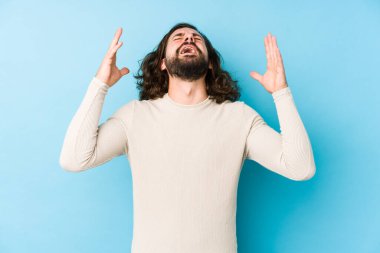 Uzun saçlı genç adam mavi bir arka planda izole edilmiş gökyüzüne haykırıyor, yukarı bakıyor, hayal kırıklığına uğramış..