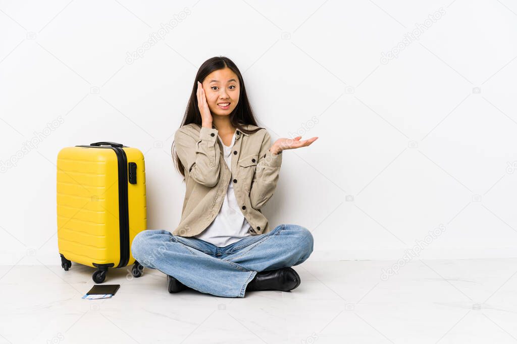 Young chinese traveler woman sitting holding a boarding passes holds copy space on a palm, keep hand over cheek. Amazed and delighted.