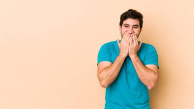 Bej arka planda izole edilmiş genç bir adam bir şeye gülüyor, elleriyle ağzını kapatıyor..