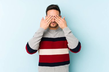 Mavi arka planda izole edilmiş genç beyaz adam elleriyle gözlerini kapatmaktan korkuyor..