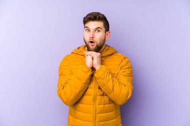 Mor arka planda izole edilmiş genç bir adam şans için dua ediyor, şaşırmış ve ağzı açık bir şekilde önüne bakıyor..