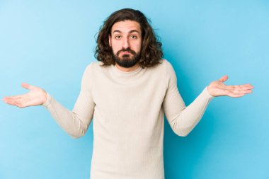 Genç, uzun saçlı, mavi bir arka planda izole edilmiş bir adam sorgulama sırasında omuz silkiyor ve kuşkulanıyor..