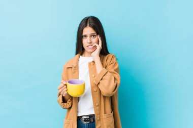 Young caucasian woman holding a coffee mug biting fingernails, nervous and very anxious.