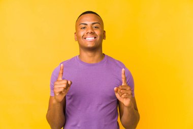 Sarı arka planda izole edilmiş genç latin adam iki ön parmağı da boş bir alan gösteriyor..