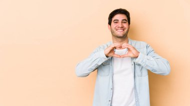 Bej arka planda izole edilmiş genç adam gülümsüyor ve elleriyle kalp şeklini gösteriyor..