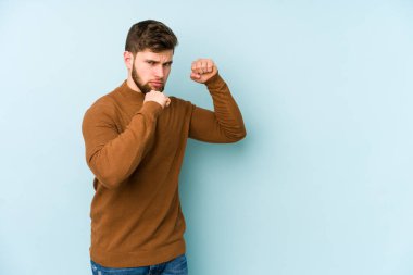 Kafkasyalı genç bir adam mavi arka planda izole edilmiş. Yumruk atıyor, sinirleniyor, kavga ediyor, kavga ediyor..