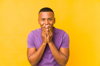 Sarı arka planda izole olmuş genç Latin bir adam bir şeye gülüyor, elleriyle ağzını kapatıyor..