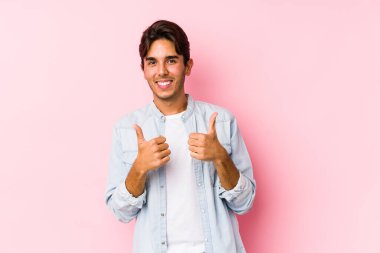 Pembe arka planda poz veren beyaz bir genç yalnız, iki parmağını da kaldırıyor, gülümsüyor ve kendinden emin..