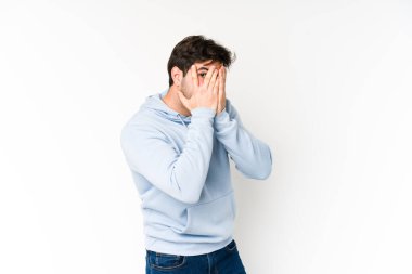 Beyaz arka planda izole edilmiş genç adam parmaklarının arasından korkmuş ve gergin bir şekilde göz kırpıyor..