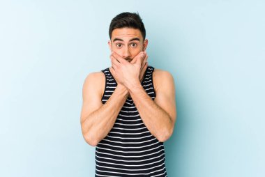 Mavi arka planda izole edilmiş genç beyaz adam elleriyle ağzını kapatırken şok oldu..