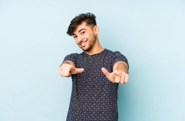 Genç Arap bir adam mavi bir arka planda soyutlanmış neşeli gülümsemeler öne doğru işaret ediyor..