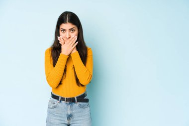 Mavi arka planda izole edilmiş genç Hintli kadın elleri endişeli bir şekilde ağzını kapatıyor..