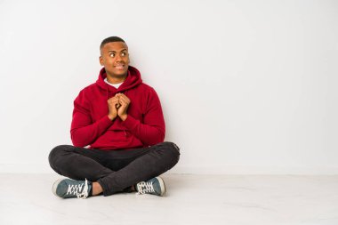 Yerde tek başına oturan genç latin adam ellerini çenesinin altında tutuyor ve mutlu bir şekilde yan tarafa bakıyor..