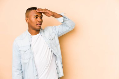 Genç latin adam bej arka planda izole edilmiş. Uzağa bakıyor. Elini alnına koyuyor..