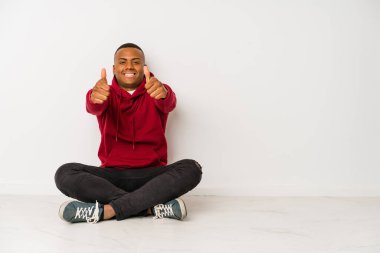 Yerde tek başına oturan genç latin adam. Baş parmakları havada, bir şey için tezahürat yapıyor. Destek ve saygı kavramı..