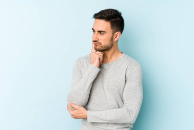 Mavi arka planda izole edilmiş genç beyaz adam şüpheli ve şüpheci bir ifadeyle yana bakıyor..