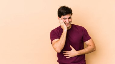 Bej arka planda izole edilmiş genç adam mutlu bir şekilde gülüyor ve ellerini karnının üstünde tutarak eğleniyor..