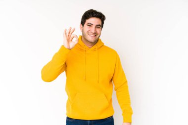 Genç adam beyaz arka planda yalnız, neşeli ve kendinden emin, iyi bir jest yapıyor..