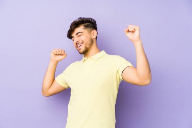 Mor arka planda izole edilmiş genç bir Arap, özel bir günü kutluyor, atlıyor ve enerjiyle kollarını kaldırıyor..