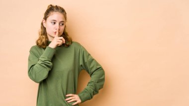 Beyazlı genç bir kadın bej arka planda izole edilmiş bir şekilde ya sır saklıyor ya da sessizlik istiyor..