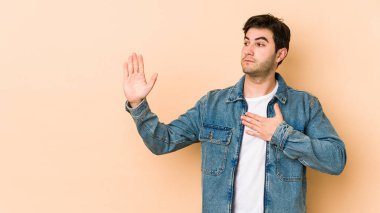 Genç adam bej arka planda izole edilmiş. Yemin ediyor, elini göğsüne koyuyor..