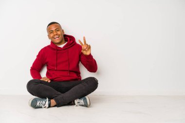 Yerde tek başına oturan genç latin adam zafer işareti gösteriyor ve gülümsüyor..